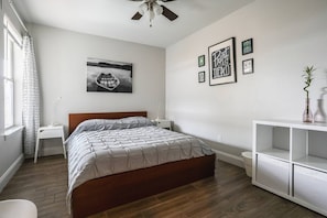 A bedroom with a wooden bed, a white nightstand, a bedside table, a white dresser, and a ceiling fan. There is a window with a patterned curtain, a framed picture on the wall, and a small potted plant on the dresser.