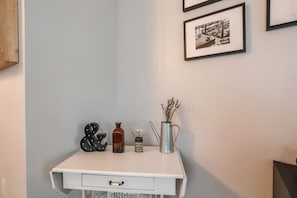 A white desk with a drawer, a vintage Edison bulb, a metal vase with dried flowers, and a black & white framed photograph on the wall.