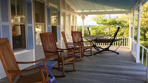 A covered porch with wooden rocking chairs and a table, white exterior walls, and a view of greenery. - Beautiful Roomy Beach House...See and Hear the Ocean Waves (Wells)