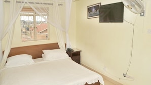 Un dormitorio con cama, mesita de noche, ventilador, televisor y ventana con vista a edificios.