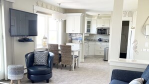 A modern living room with a dark blue sofa, a flat-screen TV, a dining area with wicker chairs, and a kitchen with white cabinets.