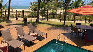 A pool area with lounge chairs and a table, surrounded by palm trees and a view of the beach. - Beachfront village with natural pools near the condominium! (Camaçari)