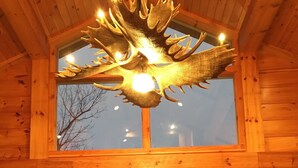 A wooden interior with a large window, a deer antler chandelier, and two green armchairs around a wooden table with a flower arrangement.