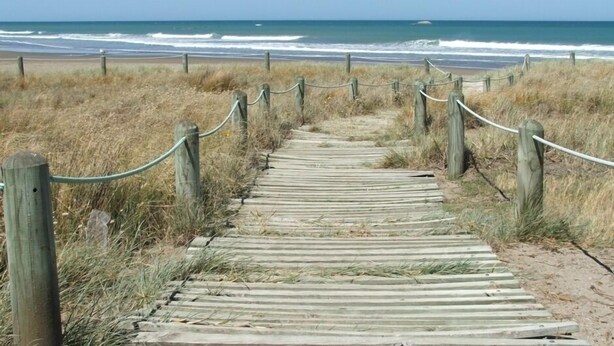 Un paseo marítimo de madera que conduce a una playa, delimitada por una valla de alambre.