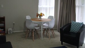 Una sala de estar con una mesa redonda, cuatro sillas, un sofá negro con un cojín estampado, un reloj en la pared y una ventana con cortinas.