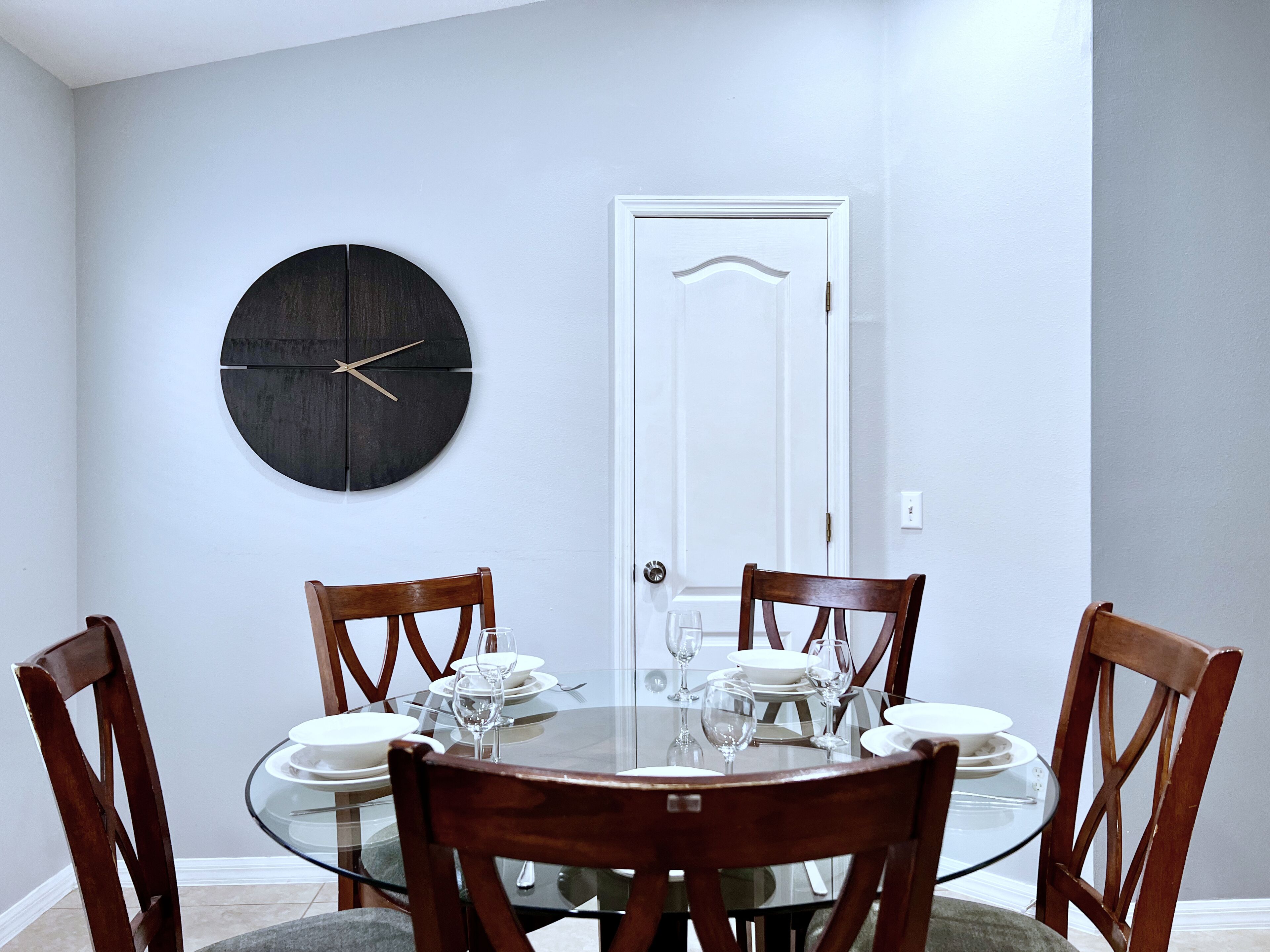 Een eetgedeelte met een ronde glazen tafel, houten stoelen, een wandklok en een witte deur.