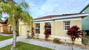 Una casa residencial con fachada amarilla, techo de tejas y un patio delantero con una palmera y diversas plantas.