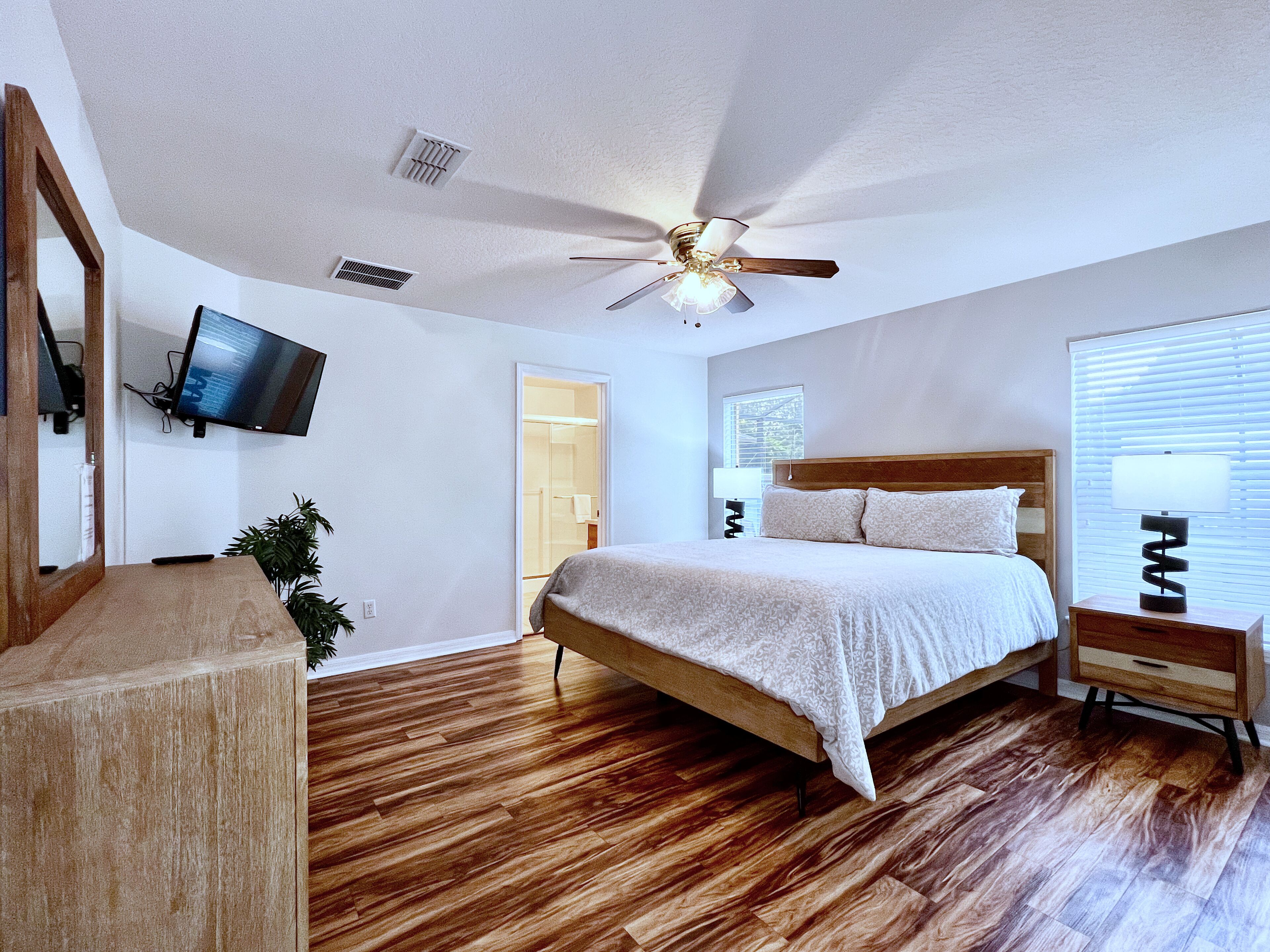 A bedroom with a wooden bed, a ceiling fan, a TV mounted on the wall, and a bedside table with a lamp.