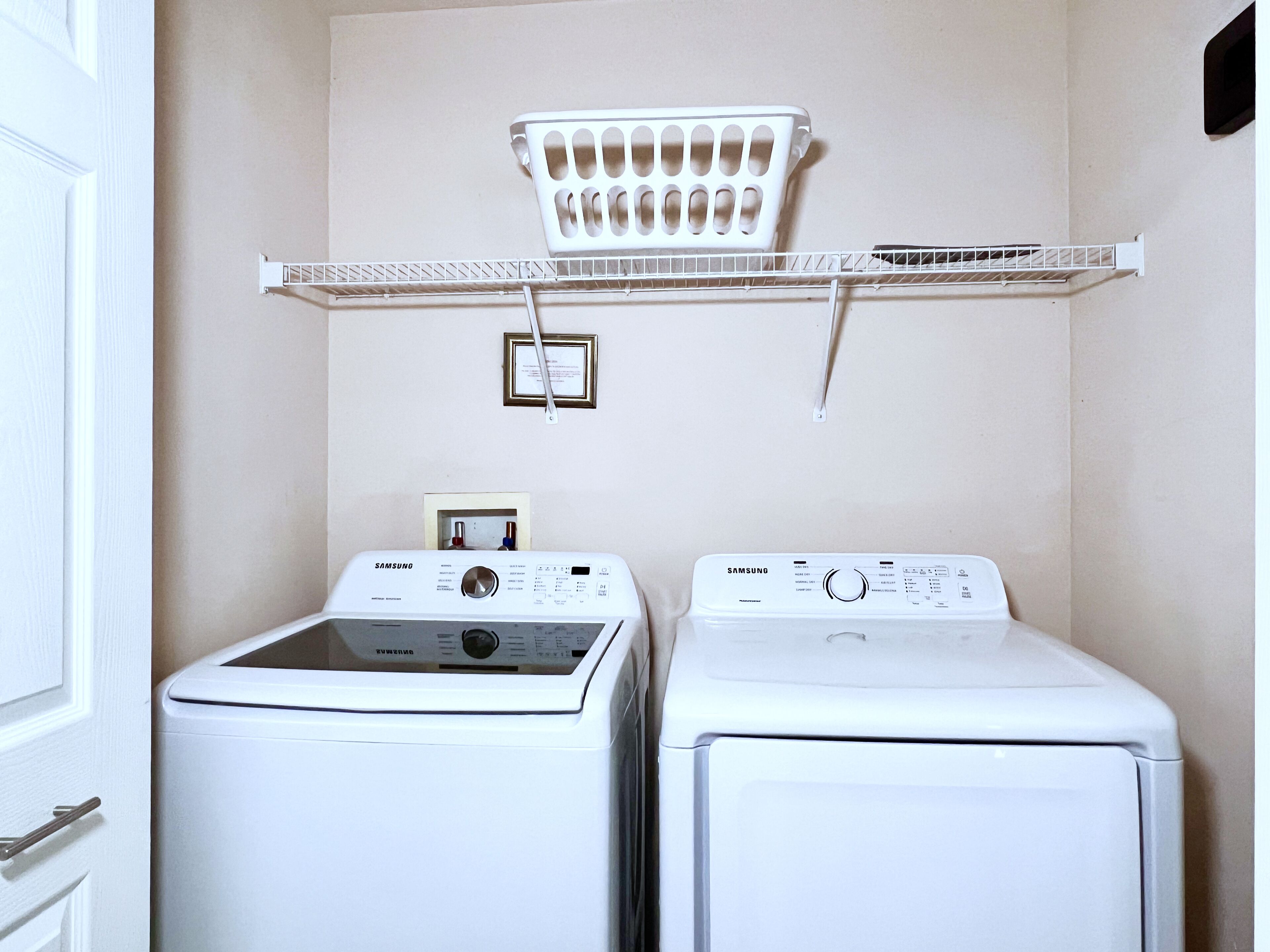 A laundry room with two white washers and dryers, a metal shelf above, and a framed certificate on the wall.