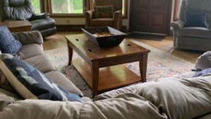 Ein Wohnzimmer mit einer Eckcouch, einem Couchtisch und zwei Sesseln. Große Fenster bieten Blick auf Grünfläche.