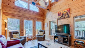A cozy cabin interior with wooden walls, large windows, a fireplace, and a television. - CREEKSIDE LOG HOME: NEXT TO CREEK / CLOSE TO TOWN / HOT TUB / TRAILS / PETS OK (Black Mountain)