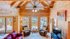 A cozy cabin interior with wooden walls, a ceiling fan, two armchairs, a red sofa, and a television. - CREEKSIDE LOG HOME: NEXT TO CREEK / CLOSE TO TOWN / HOT TUB / TRAILS / PETS OK (Black Mountain)