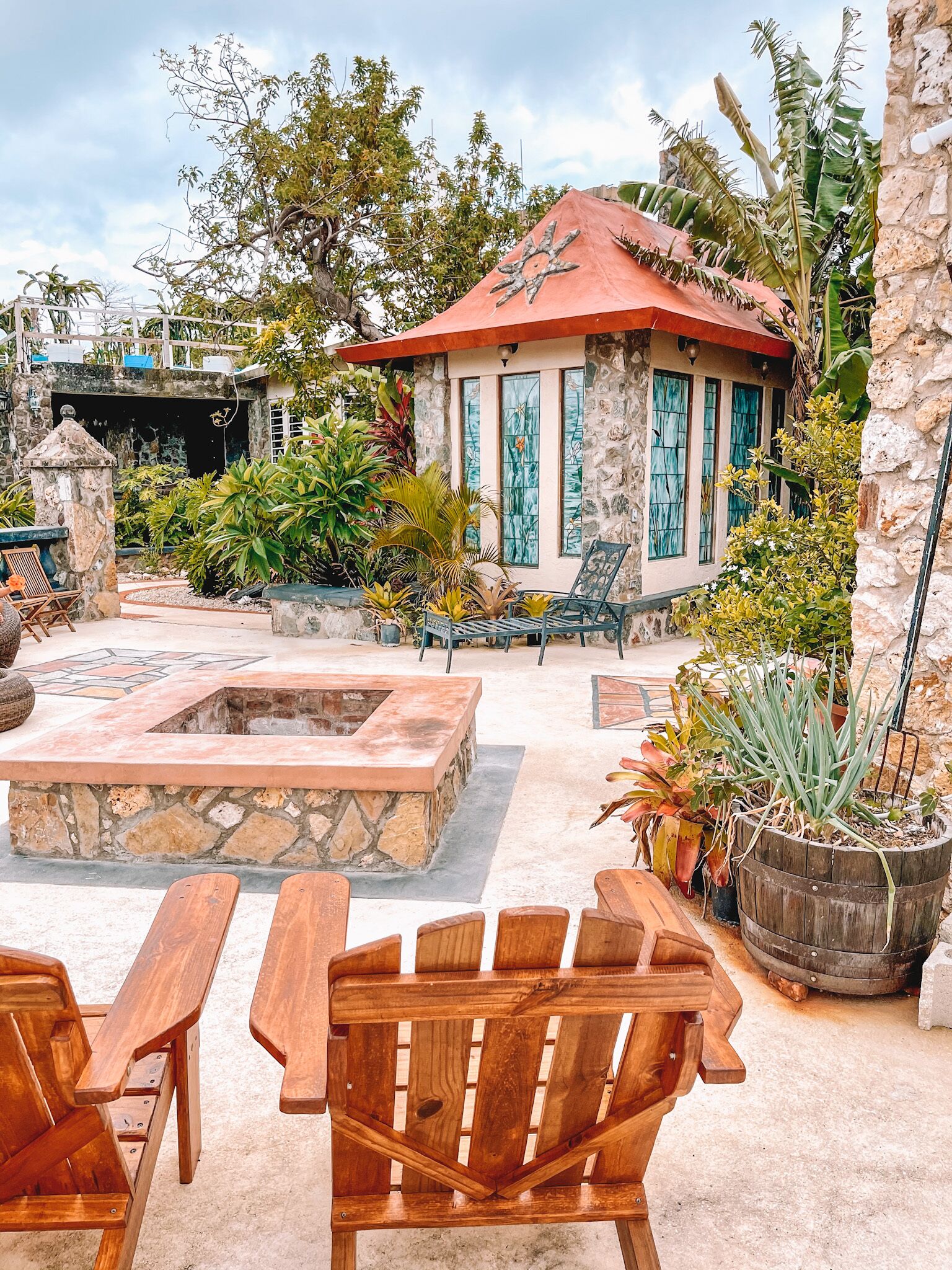 A stone building with a red roof, surrounded by tropical plants and a fire pit area with wooden benches.