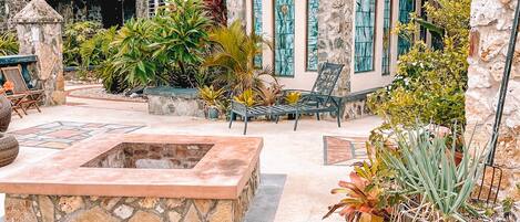 A stone building with a red roof, surrounded by tropical plants and a fire pit area with wooden benches.
