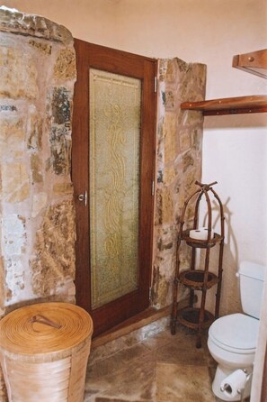 Une salle de bain avec un mur en pierre, une porte en bois ornée d’un panneau décoratif, une étagère en bois sur laquelle est posé un chandelier, et des toilettes blanches.