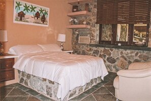 Une chambre avec un mur en pierre, un lit, une chaise, une table de chevet, une lampe, un tableau représentant des palmiers et une fenêtre avec des stores.