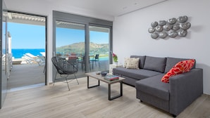 A modern living room with a grey sofa, a black chair, a coffee table, and a view of the sea through large glass doors. - Villa Gaia (Rhodes)