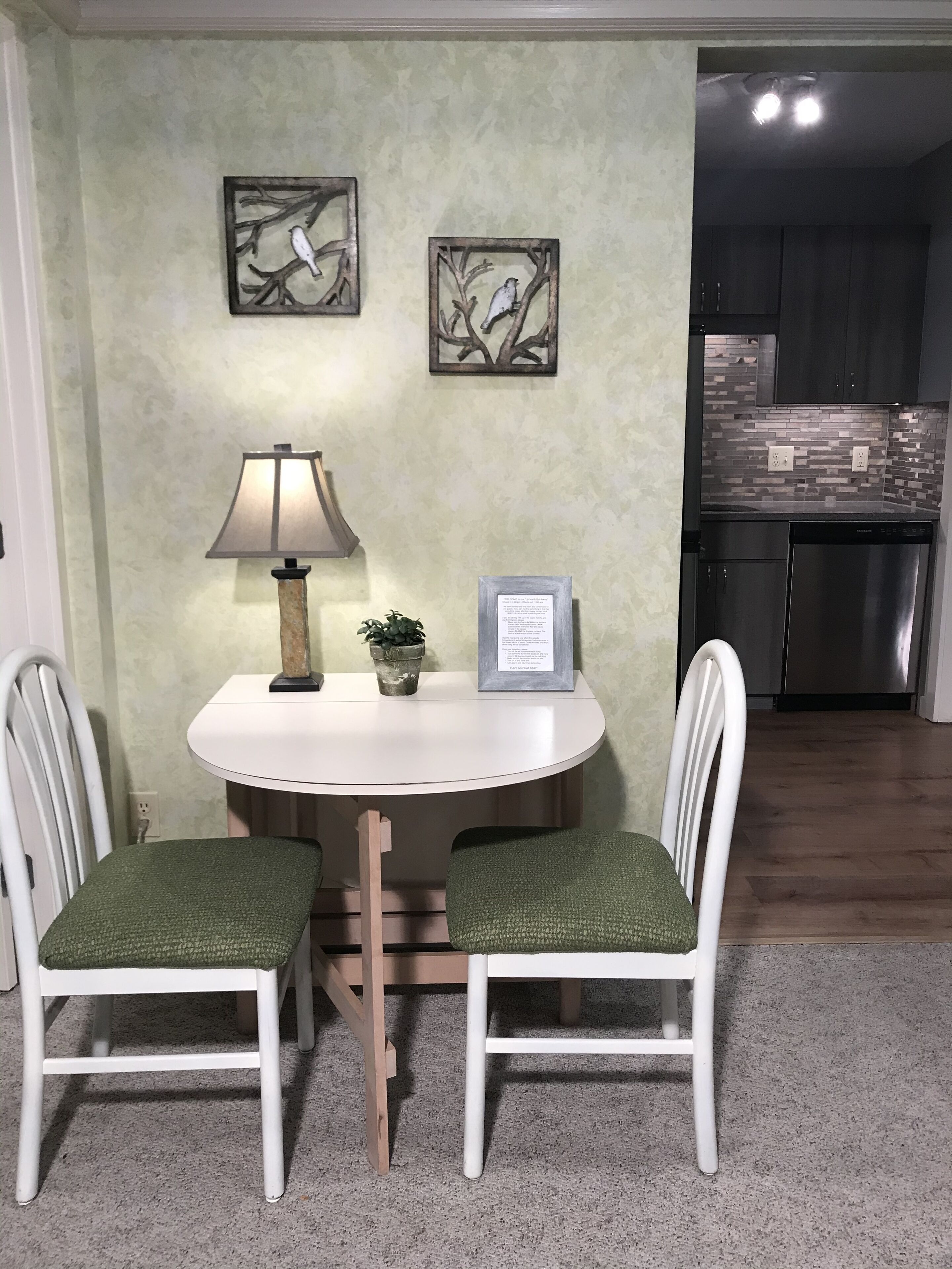 A dining area with a white table and two chairs, a lamp, a framed picture, and a small potted plant.