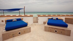 Un balcón frente a la playa con dos camas azules acolchadas, un dosel azul y vista al océano.