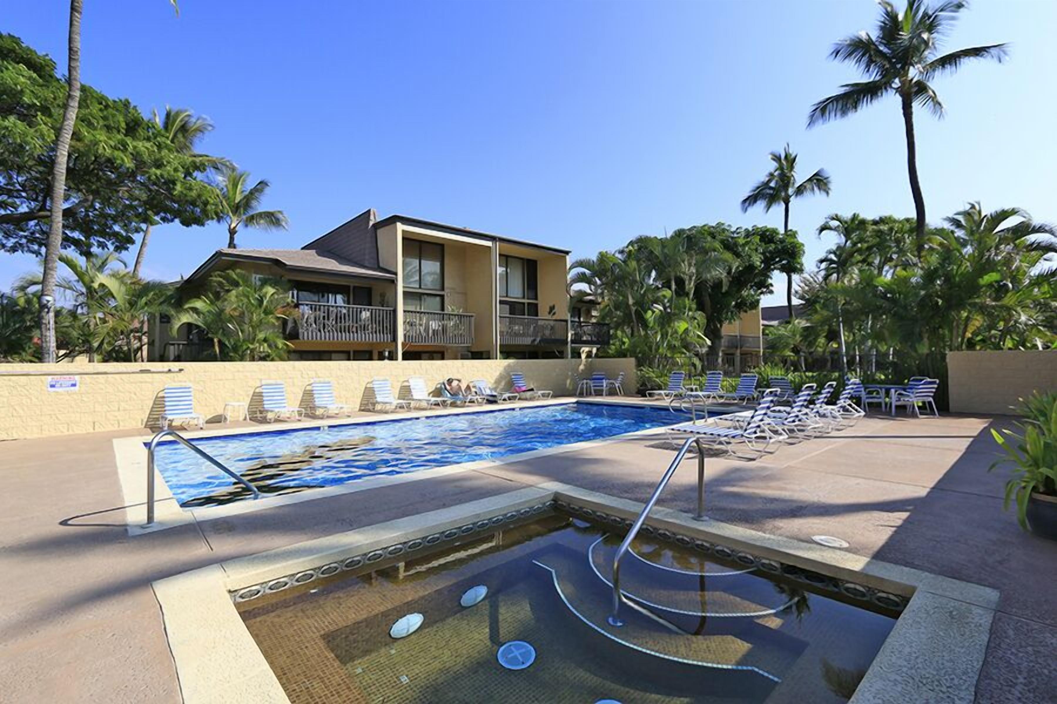 A swimming pool with lounge chairs and a hot tub, surrounded by palm trees and a building with balconies.