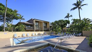 A swimming pool with lounge chairs and a hot tub, surrounded by palm trees and a building with balconies.