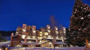 Et fleretagers hotel med snedækket grund, oplyste vinduer og et stort grantræ til højre.