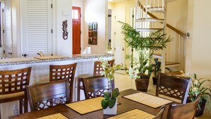 A dining area with wooden chairs and a table, a kitchen with white cabinets, and a staircase leading to an upper floor. - Quiet Top-floor Penthouse with Golf Course views, 2BR/2BA+Loft, Sleeps 6 (Puako)