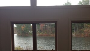 A room with a brown leather sofa, two windows with a view of a lake and trees, and a wooden floor. - Relax at Crescent Lake (wolfeboro)