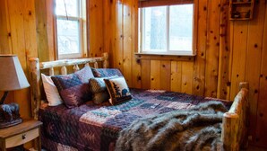 Ein Schlafzimmer in einer Holzhütte mit einem Bett, einem Nachttisch, einer Lampe und zwei Fenstern.