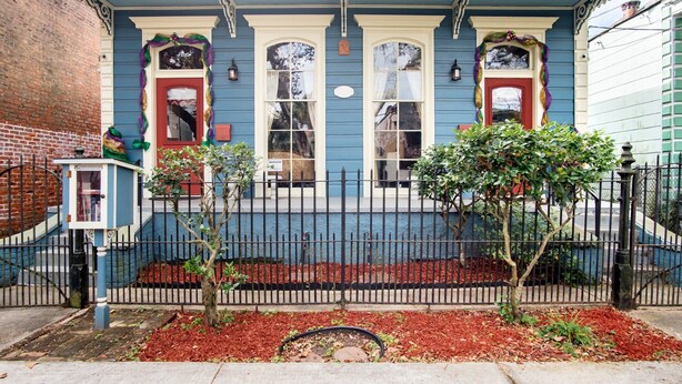 Una casa azul con detalles blancos, dos puertas rojas y un pequeño porche con barandilla.