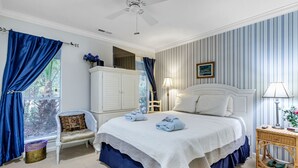 Un dormitorio con una cama grande, cabecera blanca, mesita de noche, una silla, ventana con cortinas y ventilador de techo.
