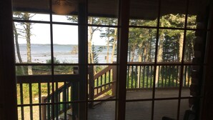 A view from a wooden deck with a railing, overlooking a beach and forested area. - Bear Beach Maine! Oceanfront Log Cabin, Secluded Sandy Beach, Sunsets & Sauna (Cutler)