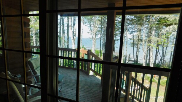 A view through a window with a wooden deck, outdoor furniture, and a railing overlooking a forested area. - Bear Beach Maine! Oceanfront Log Cabin, Secluded Sandy Beach, Sunsets & Sauna (Cutler)