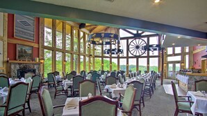Une grande salle à manger avec de hauts plafonds, de grandes fenêtres et un décor intérieur rustique en bois. Les tables sont dressées avec des nappes blanches et des chaises vertes.