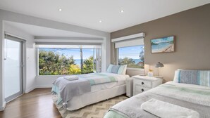 A bedroom with two beds, a large window with a view of the sea, a painting on the wall, and a bedside table with a lamp. - the nest at killcare (Killcare)