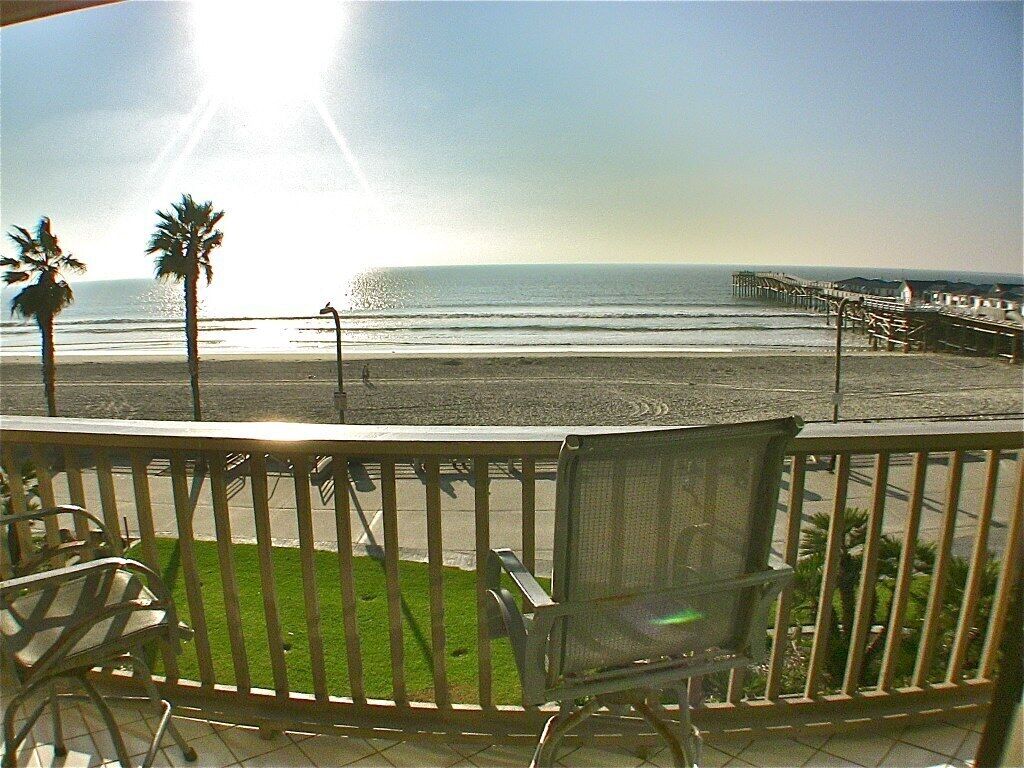 A balcony with a view of the ocean and a pier. There are chairs and a small grassy area.