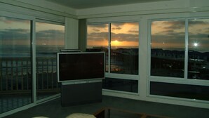 Una habitación con televisión sobre un soporte, ventanas amplias con vista al atardecer y un balcón con barandilla.