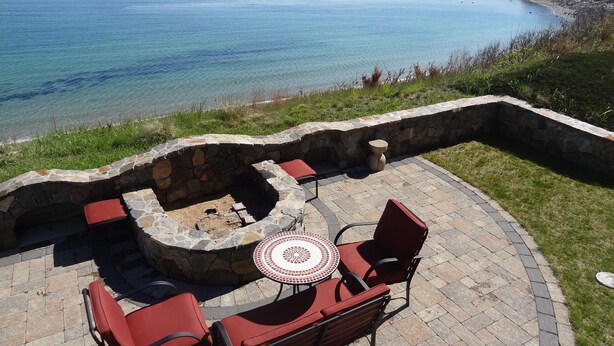 Un foyer en pierre avec des coussins rouges et une table en mosaïque, entouré de chaises rouges, offrant une vue sur la côte.