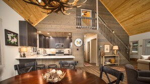 A spacious living area with a wooden ceiling, a dining table with chairs, a kitchen with dark cabinets, and a staircase leading to an upper level.