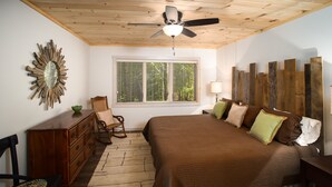 A bedroom with a wooden ceiling, a bed with a brown quilt, a wooden dresser, a chair, and a window with blinds.