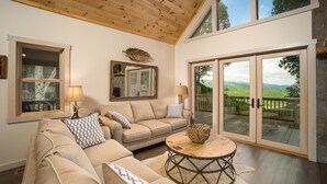 A cozy living room with a wooden ceiling, a large window, and a sliding glass door leading to a deck.