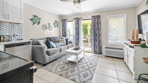 Ein modernes Wohnzimmer mit grauem Sofa, weißem Couchtisch und Deckenventilator. Es gibt eine Küchenzeile mit weißen Schränken und einem Geschirrspüler.