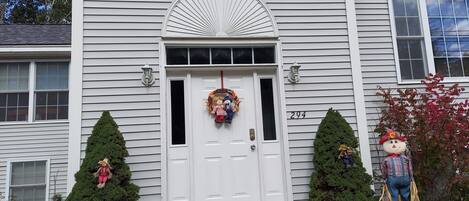 A white house with a front door, two small trees, a pumpkin, and a scarecrow decoration.