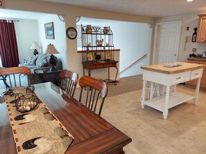 Un comedor con una mesa de madera, sillas y un tapete decorativo. Se observa también una zona de cocina con una mesa blanca y una encimera de madera.