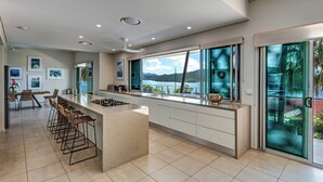 A modern kitchen with a central island, bar stools, and large windows offering a view of a lake and mountains. - Paradise@Peninsula Hamilton Island (Hamilton Island)