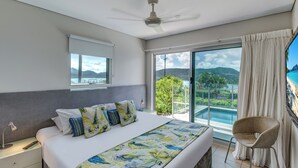 A bedroom with a large bed, a ceiling fan, a nightstand, a chair, and a view of a pool and mountains. - Paradise@Peninsula Hamilton Island (Hamilton Island)