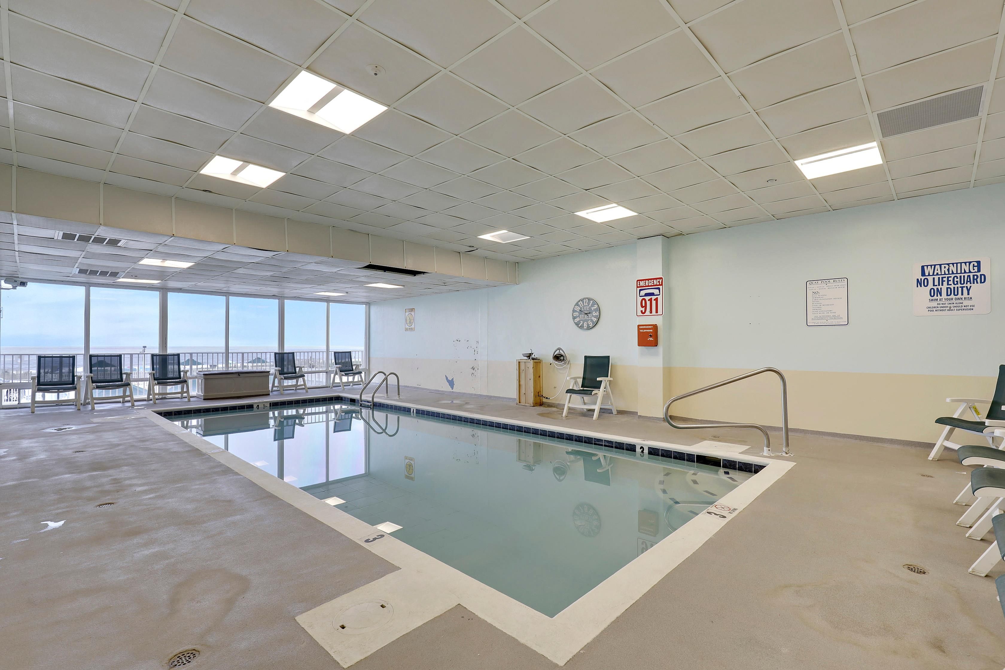 An indoor swimming pool with clear ceiling tiles, emergency exit signs, and a view of the sky through large windows.