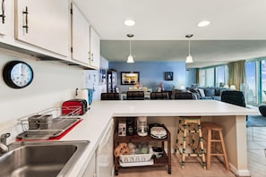 A modern kitchen with a central island, a dining area with a sofa, and a view through large windows. - XPLORIE ACTIVITIES & LINENS INCLUDED*! ! QUAY 2BR/2BA Condo with Ocean & Bay Views! (Ocean City)