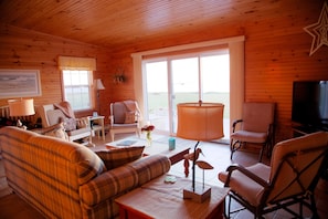 A cozy cabin interior with a plaid sofa, armchairs, a television, and a large window offering a view of the outdoors.