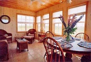 A wooden interior with a dining table set for four, surrounded by wooden chairs. There is a wooden sofa and a small table with a lamp.
